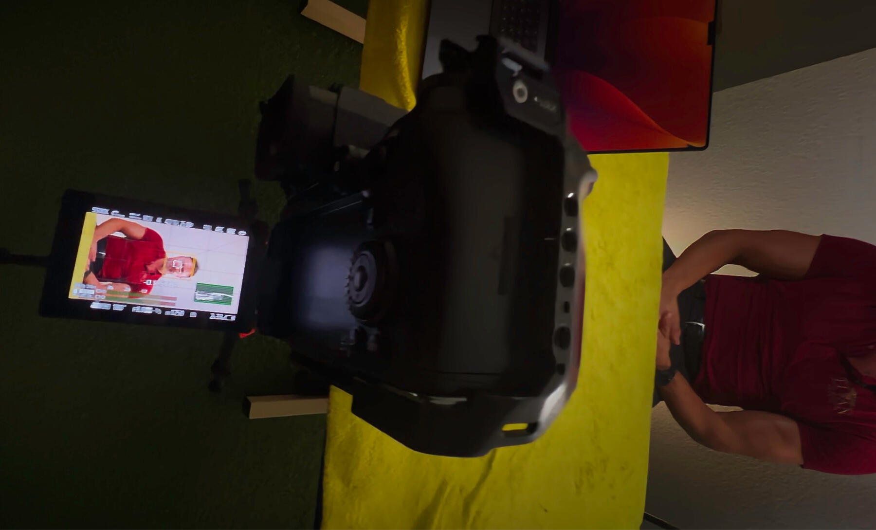 Eine Kamera auf einem Stativ filmt eine Person im roten Shirt, die auf dem Display der Kamera sichtbar ist. Auf dem Tisch davor liegt ein gelbes Tuch, daneben ein aufgeklappter Laptop.